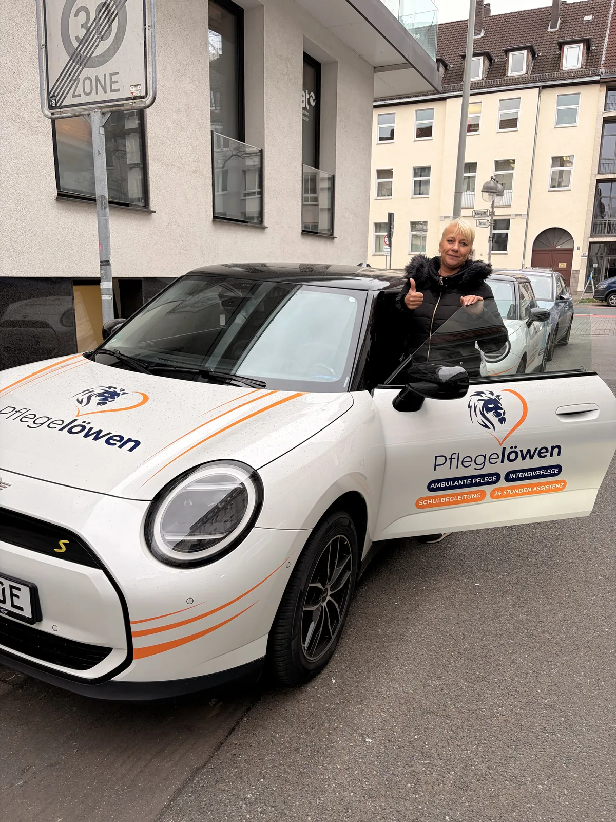 Pflegelöwen-Mitarbeiter zeigt Daumen hoch vor einem Elektroauto